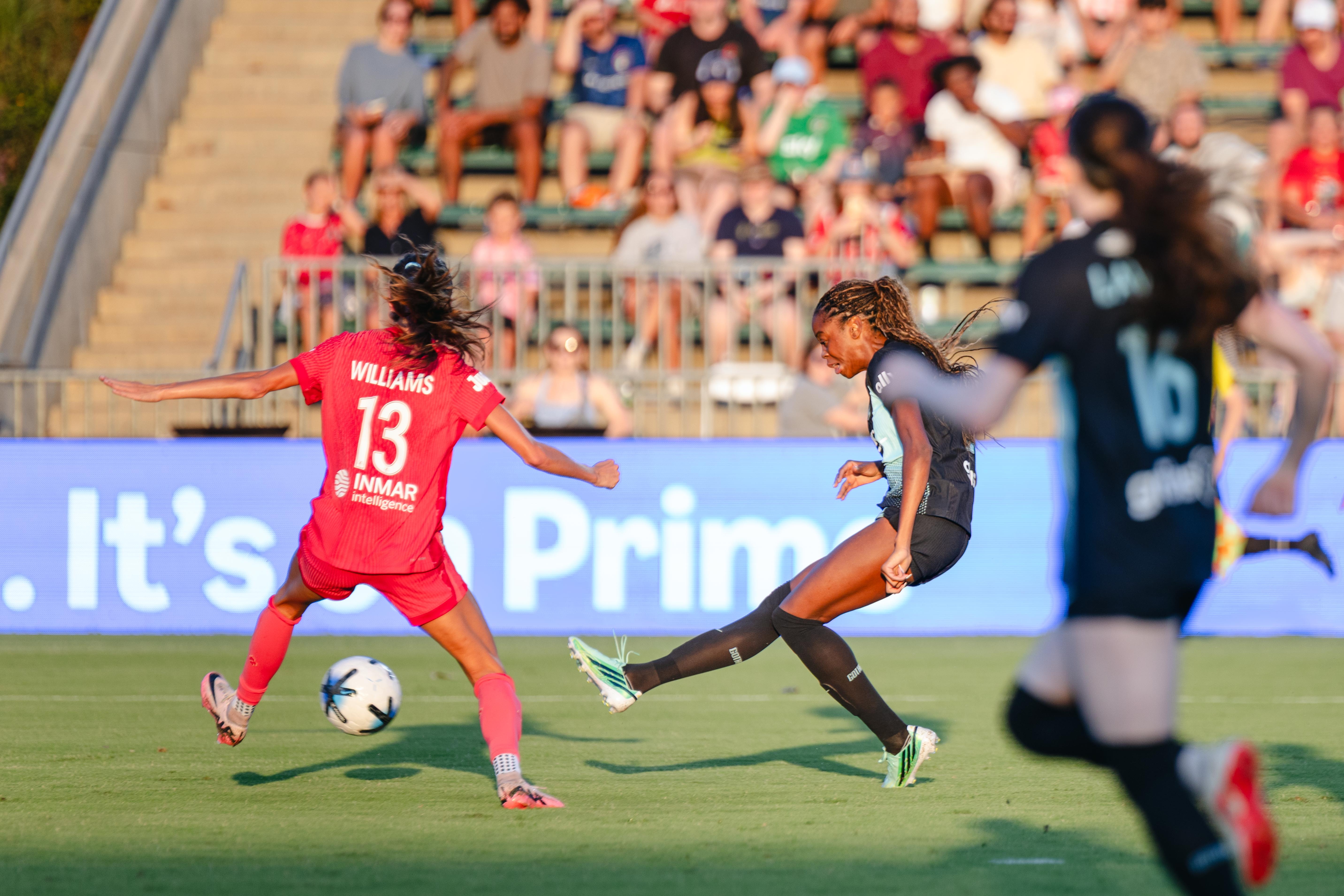 A player in a dark kit kicks the ball as a player in red moves to challenge.